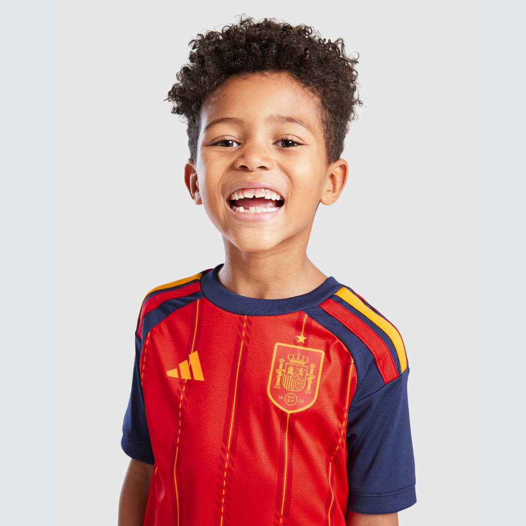 Criança vestindo a camisa infantil Espanha 2026 de frente, destacando o escudo da seleção e as listras amarelas verticais.